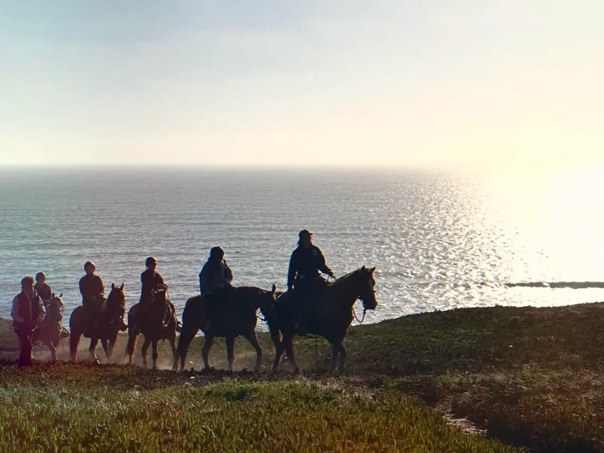 A line of riders silhouetted on the bluff above the Pacific as the low sun glitters on the water