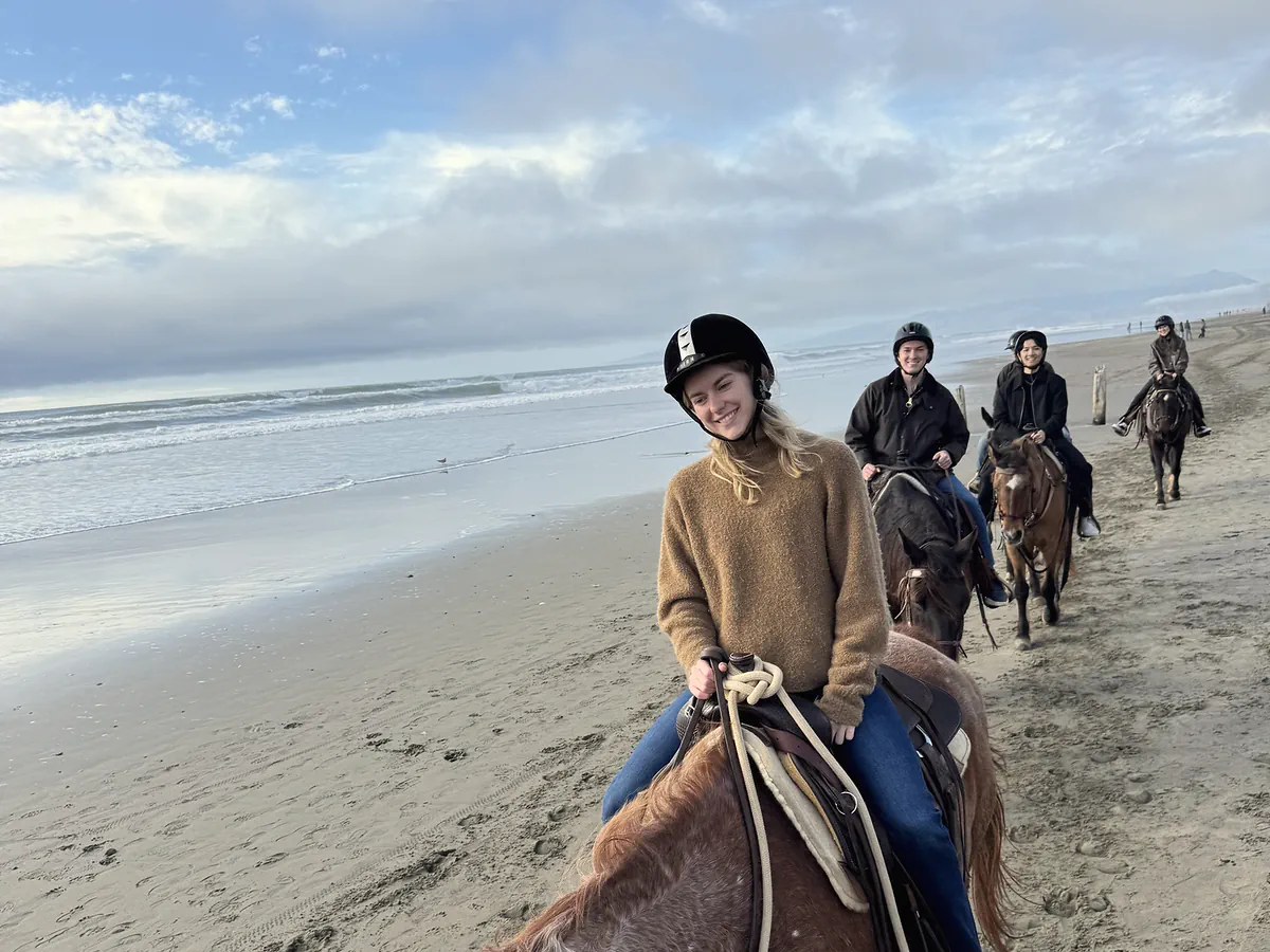 A smiling guest in a riding helmet on a chestnut roan horse on the beach, more riders in helmets behind her